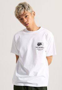 T-shirt en coton blanc avec un col rond et des manches courtes. Présente un logo noir "VANS OFF THE WALL" sur la poitrine. Coupe standard.