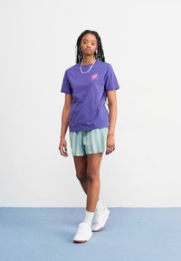Camiseta de algodón morada con cuello redondo y pequeño logo. Pantalones cortos a rayas verdes y blancas. Zapatillas blancas y calcetines completan el conjunto.