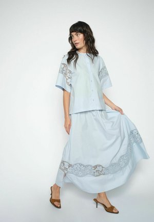 Kvinde med mørkt hår iført lyseblå knappet bluse og matchende langt skørt med blondedetaljer, poserer mod en ensfarvet hvid baggrund.