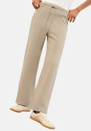 Person, der beigen, weit geschnittenen Sweatpants mit Kordelzug und hellbeigen Sneakers vor einem schlichten weißen Hintergrund trägt.