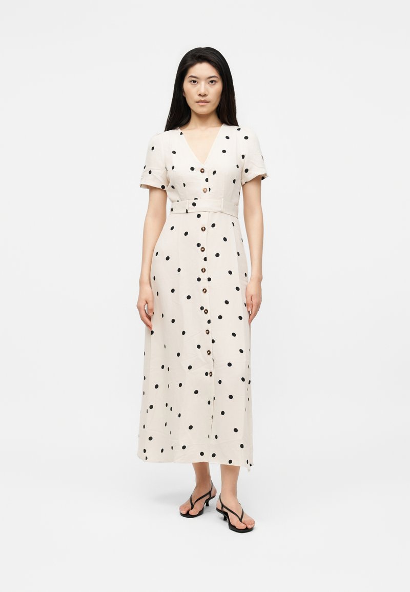 Femme portant une robe midi blanche à pois noirs avec des boutons sur le devant, associée à des sandales noires à talons.