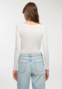 Body a maniche lunghe, aderente, in tessuto bianco, con scollatura ampia e superficie liscia, abbinato a jeans a vita alta di colore azzurro chiaro.