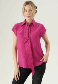 Blusa fucsia a maniche corte con fronte volants, tessuto texture e colletto, abbinata a jeans neri.