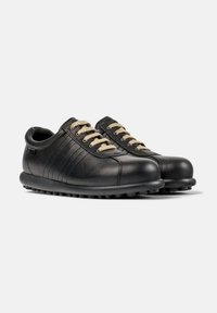 Zapatillas bajas de cuero negras con cordones beige y suelas de goma texturizada, inclinadas ligeramente hacia la derecha sobre un fondo blanco.