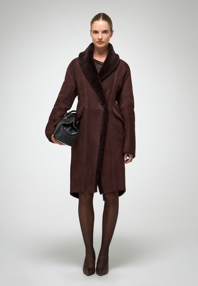 Sieviete, ģērbusies garā brūnā šermanu mētelī, melnās caurspīdīgās zeķbiksēs, brūnās papēžu kurpēs, tur melnu ādas rokassomu, fona ir vienkāršs.