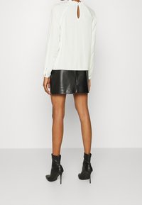 Femme portant un chemisier blanc à manches longues avec une ouverture en forme de goutte dans le dos, une mini-jupe en cuir noire et des bottines à talons hauts noires, debout.