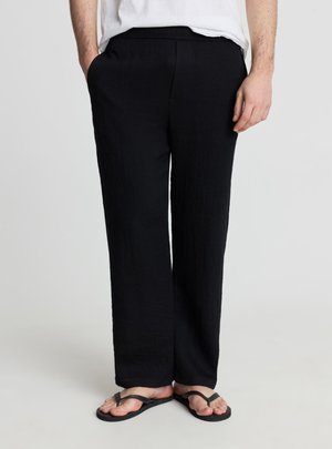 Persona che indossa pantaloni neri larghi con tasche e infradito nere, in piedi davanti a uno sfondo chiaro e uniforme.