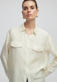 Blusa beige chiaro realizzata in tessuto testurizzato, con chiusura anteriore a bottoni, colletto a punta e due tasche sul petto. Maniche lunghe.