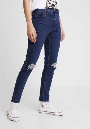 Slim-fit dark blue jeans with distressed knees worn with white high-top sneakers and a white graphic t-shirt.