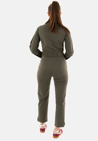 Tuta verde oliva con vita aderente e fascia elastica. Polsini con dettagli a bottone alle caviglie. Indossata con scarpe bianche e bordeaux.