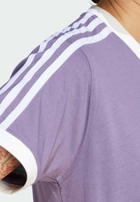 Lila idrottströja med vita detaljer och tre vita ränder på axeln. Mjuk tyg med korta ärmar och v-ringning.
