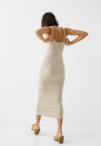 Kvinna med axellångt mörkt hår som justerar remmarna på en åtsittande beige midiklänning, iförd beigefärgade klackskor, st ående mot en enkel vit bakgrund.