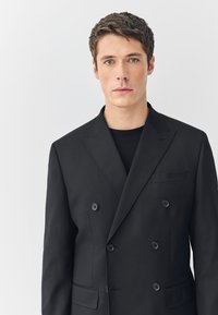 Fekete, kétsoros, csúcsos hajtókás blézer, két első zsebbel és hat gombbal. Az anyag sima, ropogós textúrájú.