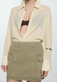 Femme portant une chemise beige boutonnée, partiellement ouverte pour montrer un soutien-gorge marron, et une mini-jupe kaki avec des poches à l'avant.