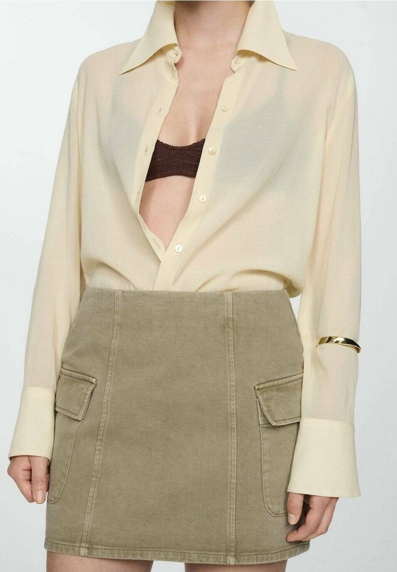 Femme portant une chemise beige boutonnée, partiellement ouverte pour montrer un soutien-gorge marron, et une mini-jupe kaki avec des poches à l'avant.
