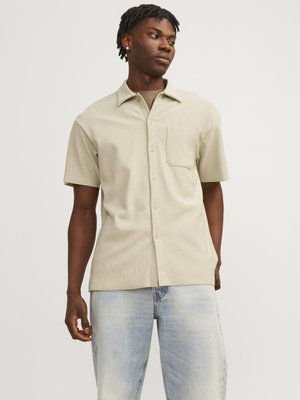 Chemise à manches courtes de couleur beige clair avec un col, une poche poitrine unique et une texture côtelée subtile, associée à un jean en denim bleu clair.