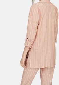 Chemise longue rose clair avec des rayures blanches verticales, manches retroussées, coupe décontractée et ourlet carré.