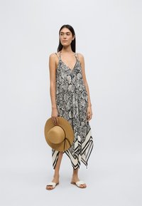 Μακριά ασπρόμαυρη φορεμα με μοτίβο, V-λαιμόκοψη, λεπτές τιράντες και ασύμμετρο τελείωμα· συνοδευόμενη από ψάθινο καπέλο με φαρδύ γείσο.