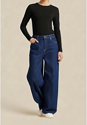 Woman wearing black long-sleeve top tucked into high-waisted wide-leg blue jeans with white shoes, standing with one hand in pocket.