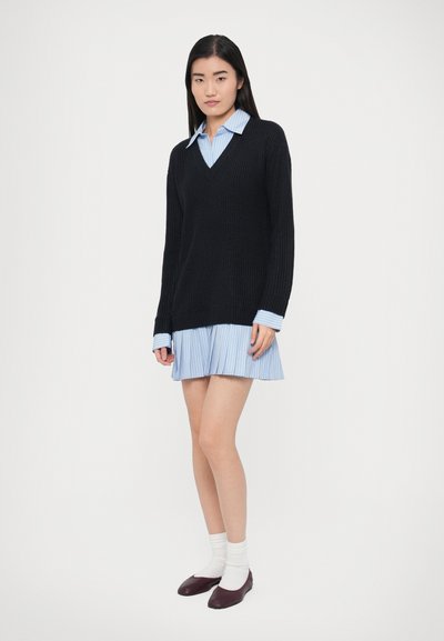 Fekete, V-nyakú kötött pulóver, világoskék pliszírozott ruha fölött rétegezve. A modell fehér zoknit és bordó balerinacipőt visel.