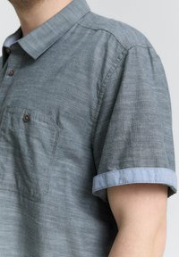 Korte mouw blouse van een gestructureerde grijze stof met een lichtblauwe kraag en opgerolde cuffs. Heeft een opgestikte zak en bruine knopen.