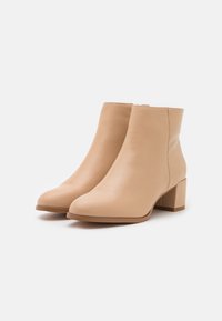 Par de botas de cuero beige hasta el tobillo con tacones cuadrados y cremalleras laterales sobre un fondo blanco.