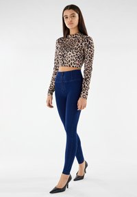 Crop top a maniche lunghe con stampa leopardata abbinato a jeans skinny aderenti blu scuro a vita alta e tacchi a spillo neri, su uno sfondo neutro.