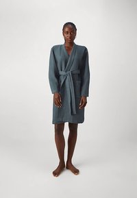 Dunkelgrüner Bademantel mit Waffelstruktur, langen Ärmeln, Gürtel und knielangem Schnitt. Barfußes Model steht vor neutralem Hintergrund.