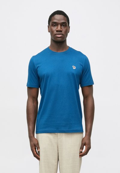 Niebieska koszulka z krótkim rękawem wykonana z bawełny, z małym haftowanym wzorem na lewej piersi. Gładka faktura, okrągły dekolt.