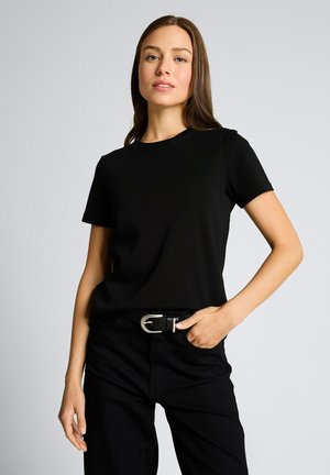 Jeune femme aux longs cheveux bruns portant un t-shirt noir uni, un pantalon noir et une ceinture noire avec une boucle argentée, debout devant un fond gris.