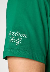 Tricou scurt cu mânecă verde, cu o textură moale. Prezintă un text brodat "Malbon Golf" în alb pe mâneca stângă.
