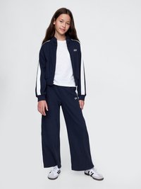 Navy tracksuit with white side stripes, featuring a zip-up jacket and wide-leg pants. White t-shirt underneath, paired with white and black sneakers.