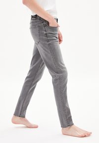 Grey denim jeans with a slim fit and mid-rise. Features five pockets, belt loops, and subtle fading. Worn barefoot, displayed in profile.