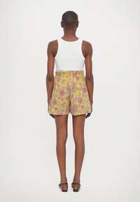 Pantaloni corti floreali gialli con motivi rosa e viola, design a vita alta, parte anteriore plissettata e tasche laterali. Abbinati a una canotta bianca.