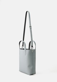 Sac fourre-tout en cuir gris avec une sangle noire ajustable, au design élancé, deux poignées supérieures et une texture lisse. Structure minimaliste et accents en métal argenté.
