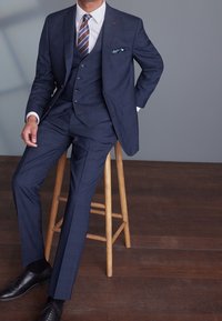 Marineblau karierter Dreiteiler mit einem weißen Hemd, gestreifter Krawatte und Einstecktuch. Schwarze Lederschuhe und ein Holzstuhl runden den Look ab.