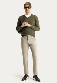 Suéter verde oliva de cuello en V, pantalones beige, zapatillas deportivas negras. Bolsa marrón de hombro. La modelo está de pie con las manos entrelazadas, llevando gafas de sol.