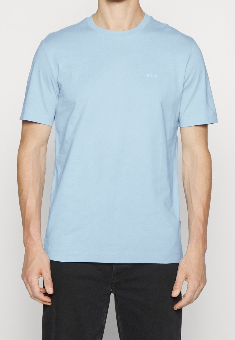 Homme portant un t-shirt bleu clair uni à manches courtes et un pantalon noir, debout devant un fond uni.