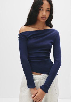 Top lungo blu navy a spalle scoperte in tessuto morbido con dettagli arricciati in vita, abbinato a pantaloni della tuta grigi chiari.
