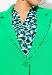 Světle zelený blazer s jedním knoflíkem, přes kterým je tričko s vzorem v černé, modré a zelené kruhové motivy. Hladká textilie.