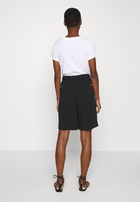Pantaloni corti neri con vita elastica e tasche laterali, abbinati a una t-shirt bianca semplice, visti da dietro. Tessuto dalla texture liscia.