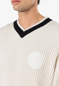 Cream-colored knitted sweater with black ribbed V-neck and a raised circular logo on the chest, showcasing a textured stitch pattern.