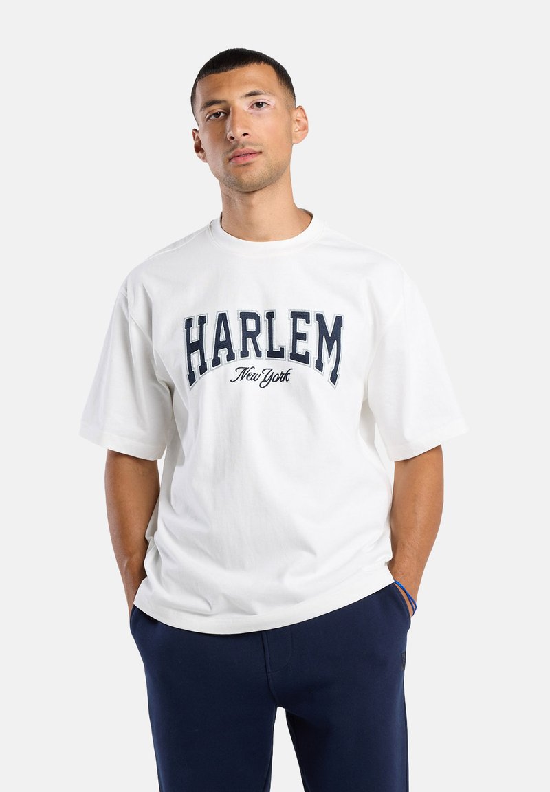 Man staat met handen in zijn zakken, draagt een wit "Harlem New York" T-shirt en donkerblauwe broek tegen een effen achtergrond.