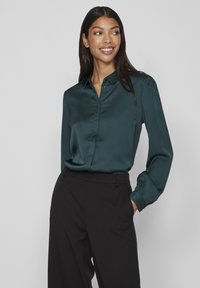 Blusa in satin turchese con colletto a delle bottoni, maniche lunghe e polsini arricciati, abbinata a pantaloni neri a vita alta. Texture liscia.