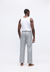 Vīrietis stāv basām kājām, ar muguru pret skatītāju, ģērbies baltā bez piedurknēm krekliņā un pelēkām un baltām vertikālām svītrām biksēs ar logotipa jostu.