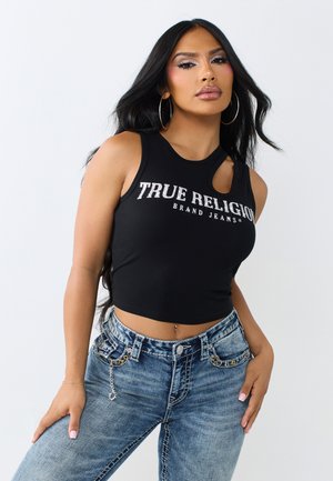 Vrouw met lang zwart haar, die hoepeloorbellen draagt, een zwart crop top met de tekst "True Religion" en blauwe jeans met kettingdetails.