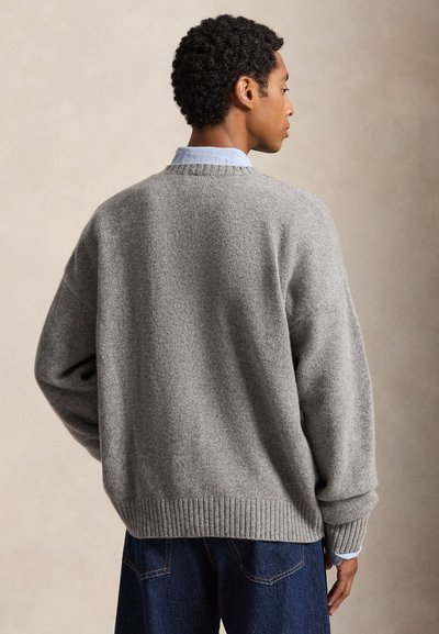 Homme vu de dos portant un pull gris ample par-dessus une chemise bleu clair et un jean bleu foncé sur un fond beige uni.