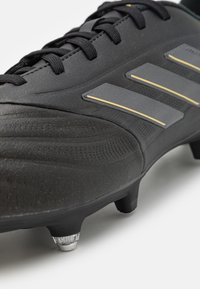 Botín de fútbol negro con parte superior de cuero texturizado, que presenta tres franjas grises y detalles en dorado. Punta redondeada y tacos de metal en la suela.