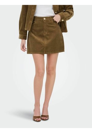 Femme portant une mini-jupe en velours côtelé marron, une veste assortie, un top blanc et des talons marron ouverts à l'avant, debout avec une main dans la poche.
