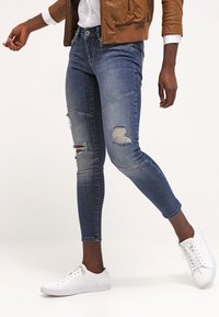 Jeans ajustados, desgastados en azul con detalles de parches, con una cintura de altura media, largos hasta el tobillo, combinados con zapatillas blancas.